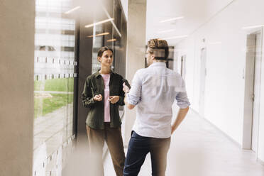 Young businesswoman talking to colleague standing in office corridor - JOSEF23398
