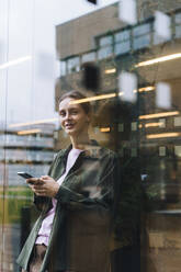 Smiling young businesswoman holding smart phone and leaning on glass - JOSEF23412