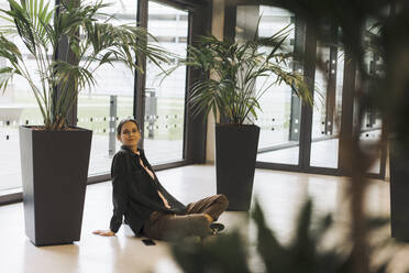 Smiling businesswoman sitting near plants on floor - JOSEF23414