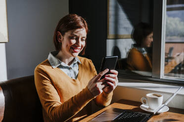 Smiling freelancer using smart phone and sitting with laptop in cafe - EBBF08440