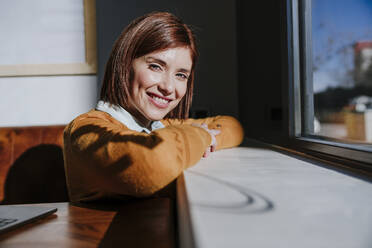 Smiling woman leaning on window sill - EBBF08445