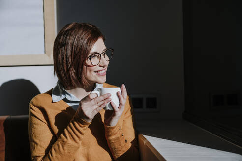 Lächelnde Frau hält Kaffeetasse und genießt das Sonnenlicht - EBBF08450