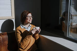 Lächelnde Frau hält Kaffeetasse und genießt das Sonnenlicht in einem Café - EBBF08452