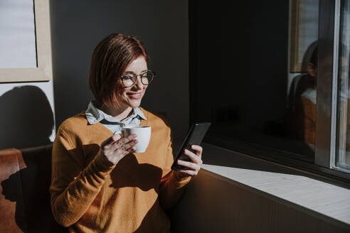 Lächelnde Frau hält eine Kaffeetasse und benutzt ein Smartphone in einem Café - EBBF08453