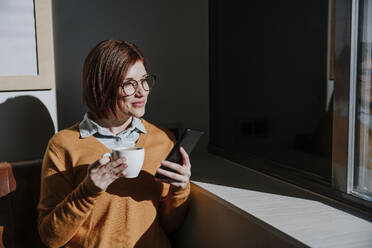 Happy woman holding coffee cup and smart phone in cafe - EBBF08454