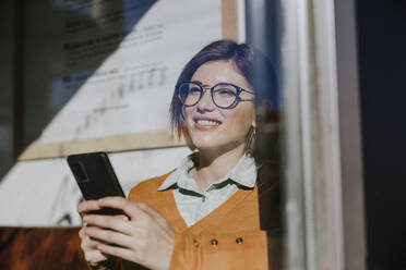 Glückliche Frau mit Smartphone durch Glas gesehen - EBBF08456