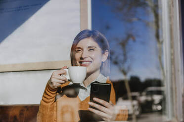 Glückliche Frau sitzt mit Smartphone und Kaffeetasse in einem Café - EBBF08457