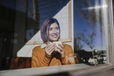 Glückliche Frau sitzt mit Kaffeetasse in einem Café - EBBF08458