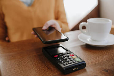 Woman paying through smart phone on credit card reader near coffee cup at table - EBBF08462