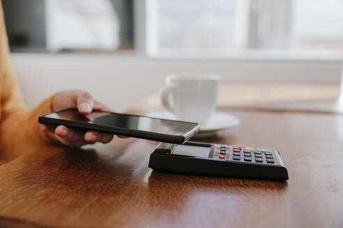 Hand einer Frau, die mit ihrem Smartphone auf einem Kreditkartenlesegerät neben einer Kaffeetasse am Tisch bezahlt - EBBF08463