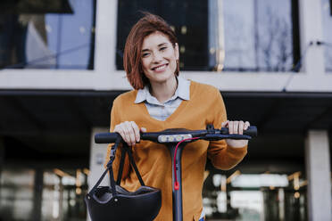 Lächelnde Frau mit Elektro-Scooter vor einem Gebäude stehend - EBBF08466