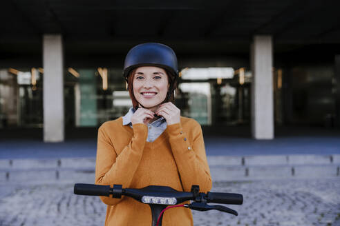 Lächelnde Frau passt ihren Helm an und steht in der Nähe eines Gebäudes - EBBF08469