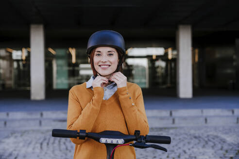 Smiling woman adjusting helmet and standing near building - EBBF08469