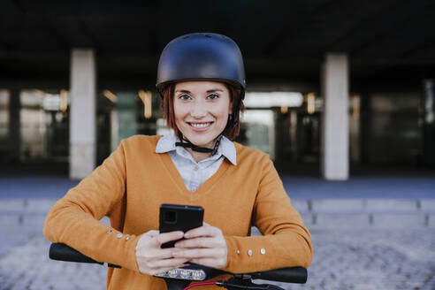 Smiling woman standing with smart phone near building - EBBF08470