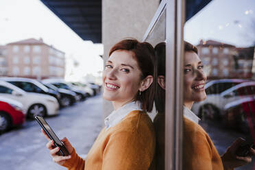 Lächelnde Frau lehnt mit Smartphone an einem Glas - EBBF08473