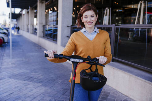 Happy woman standing with electric push scooter at footpath - EBBF08478