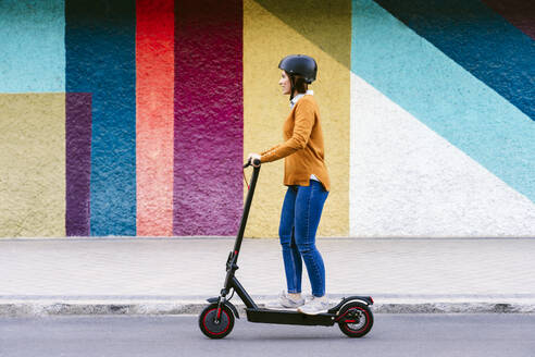 Woman wearing helmet and riding electric push scooter in front of multi colored wall - EBBF08490