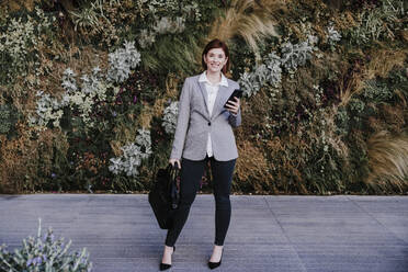 Glückliche Geschäftsfrau mit Tasche und Smartphone vor Pflanzen stehend - EBBF08492