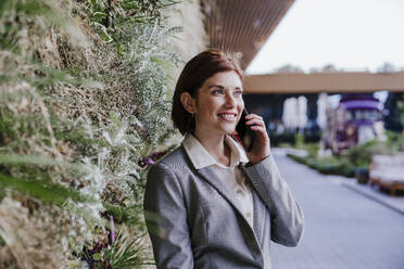 Happy businesswoman talking on smart phone near plants - EBBF08493