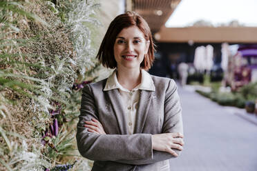 Happy businesswoman standing with arms crossed near plants - EBBF08495