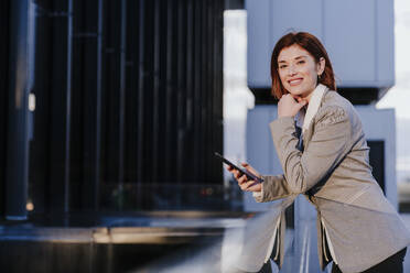 Lächelnde Geschäftsfrau steht mit Smartphone in der Nähe von Gebäuden - EBBF08500