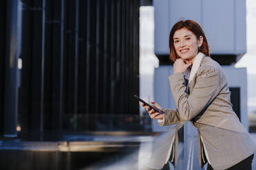 Smiling businesswoman standing with smart phone near buildings - EBBF08500