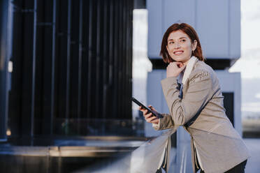 Happy businesswoman standing with smart phone near buildings - EBBF08501