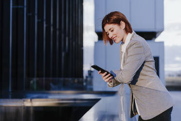 Happy businesswoman using smart phone near buildings - EBBF08502