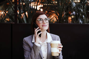 Businesswoman holding coffee cup and talking on smart phone in cafe - EBBF08512