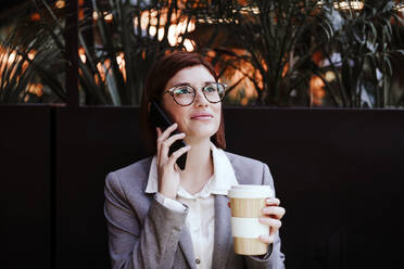 Geschäftsfrau hält Kaffeetasse und spricht mit Smartphone im Café - EBBF08512