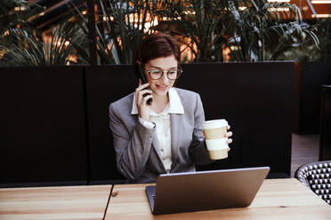 Lächelnde Geschäftsfrau, die eine Kaffeetasse hält und mit einem Smartphone in einem Café spricht - EBBF08513