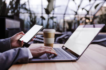 Geschäftsfrau hält Smartphone und tippt auf Laptop in Café - EBBF08515