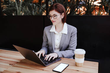 Smiling businesswoman working on laptop at table in cafeteria - EBBF08518