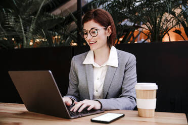 Glückliche Geschäftsfrau arbeitet am Laptop am Tisch in der Büro-Cafeteria - EBBF08519