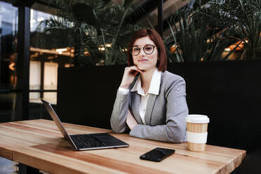 Lächelnde Geschäftsfrau sitzt mit Laptop und Smartphone in der Nähe einer Kaffeetasse am Tisch - EBBF08521