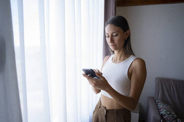 Junge Frau benutzt Smartphone in der Nähe des Fensters zu Hause - FBAF02124