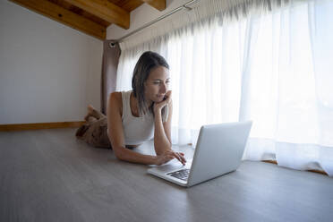 Freelancer lying on floor and using laptop near window at home office - FBAF02131