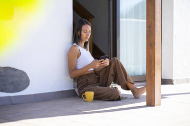 Young woman using smart and sitting on front porch - FBAF02142