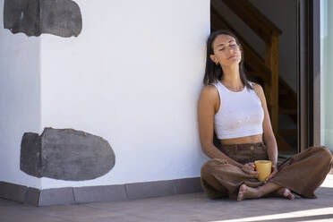 Woman holding mug and sitting with eyes closed at front porch - FBAF02143