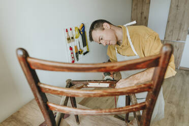 Man repairing old wooden chair using sander tool at home - YTF01771