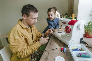 Tochter schaut auf Vater, der zu Hause an der Nähmaschine die Nadel mit einer Schere schneidet - YTF01782