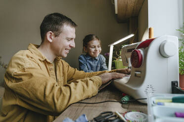 Happy daughter with father sewing recycled clothes through machine at home - YTF01792