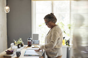 Side view of woman examining financial bills while standing in kitchen at home - MASF42240