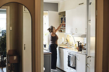 Side view of mature woman with disability searching in cabinet while standing in kitchen at home - MASF42245