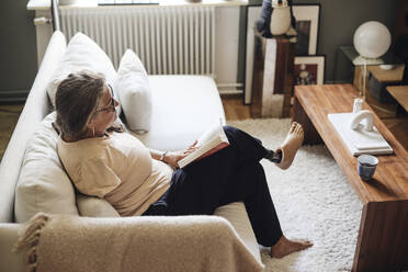 High angle view of woman with disability reading book while sitting on sofa at home - MASF42249