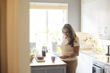 Mature woman using smart phone while standing near kitchen island at home - MASF42276