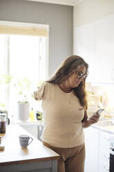 Mature woman with disability surfing net through smart phone while standing in kitchen at home - MASF42284