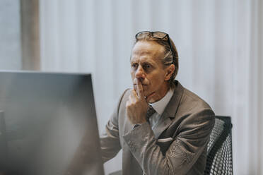 Reifer Geschäftsmann mit Hand am Kinn, der auf einen Computerbildschirm im Büro schaut - MASF42292