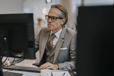 Mature businessman wearing eyeglasses using computer at desk in office - MASF42297