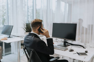 Rückansicht eines Geschäftsmannes, der am Computerschreibtisch im Büro sitzt und mit einem Mobiltelefon spricht - MASF42312